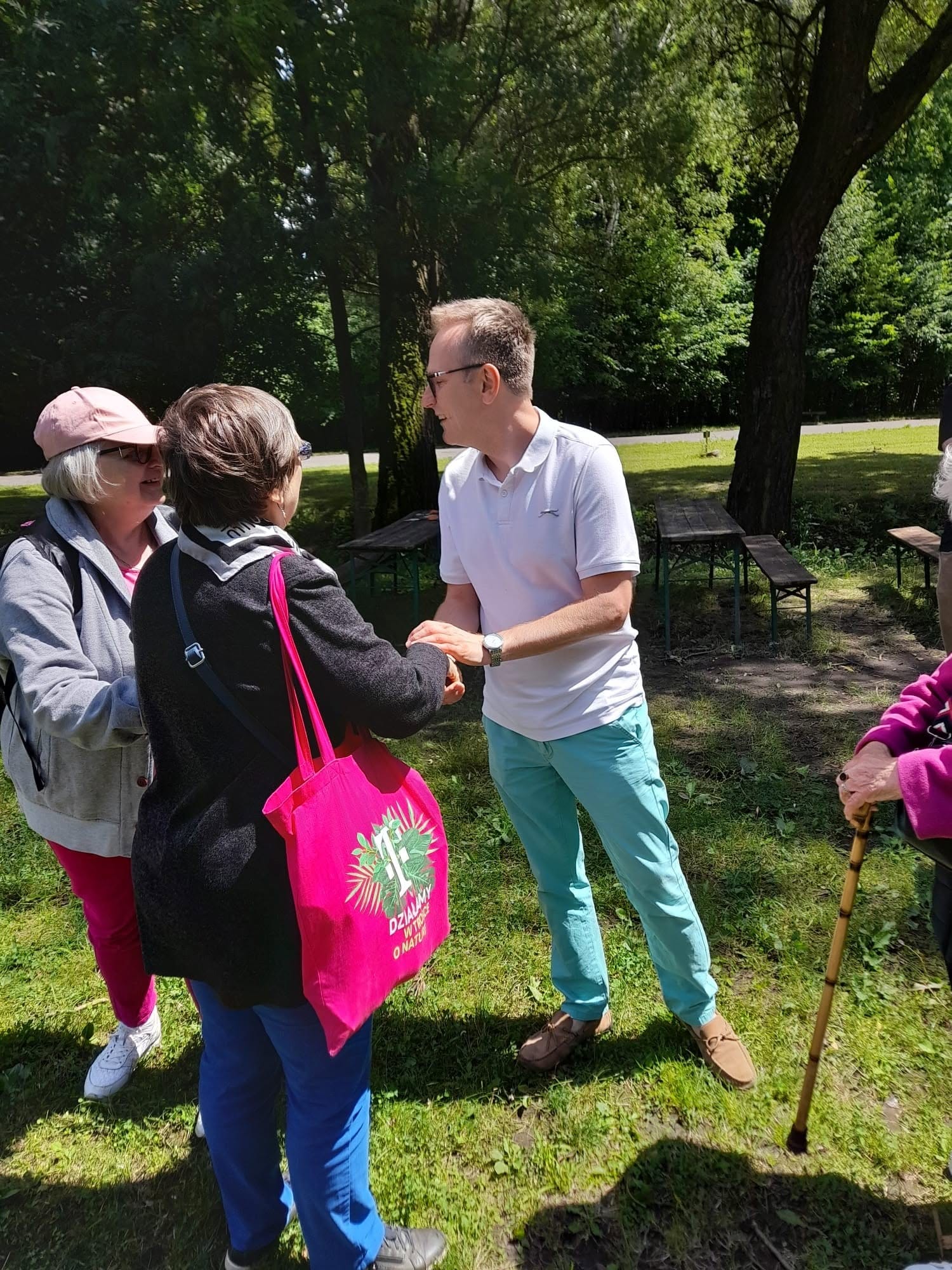 Mężczyzna witający się uściskiem dłoni z uczestniczkami wycieczki w parku na świeżym powietrzu.