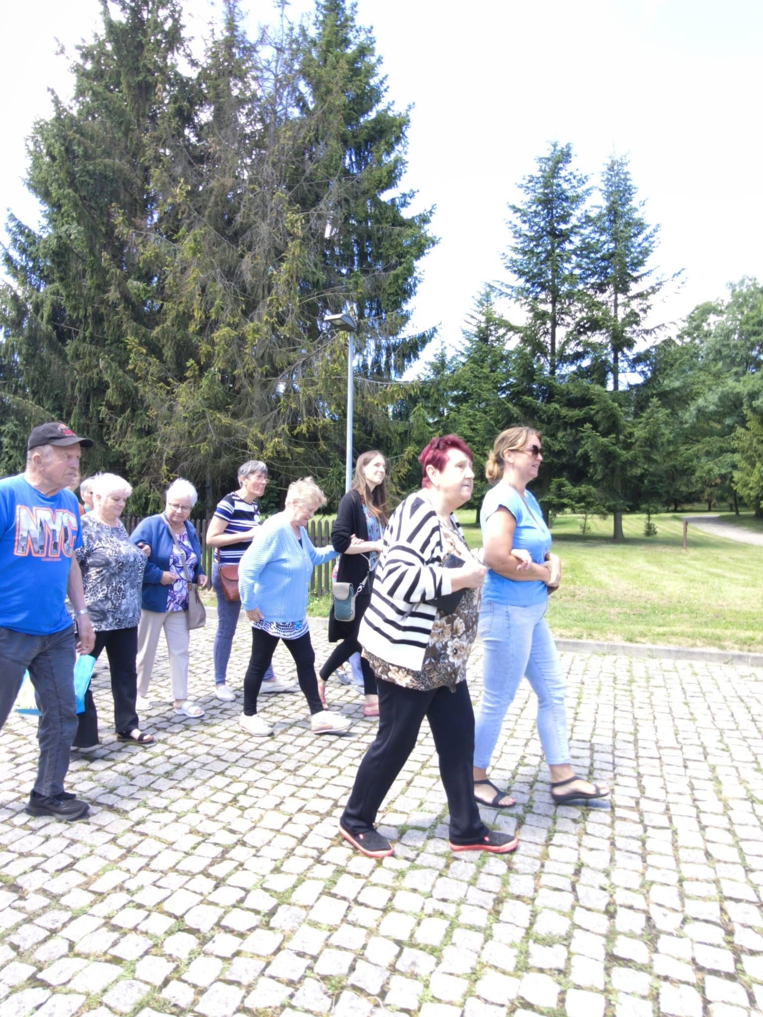 Grupa osób spacerująca z uśmiechami po brukowej ścieżce w otoczeniu wysokich drzew i zieleni.