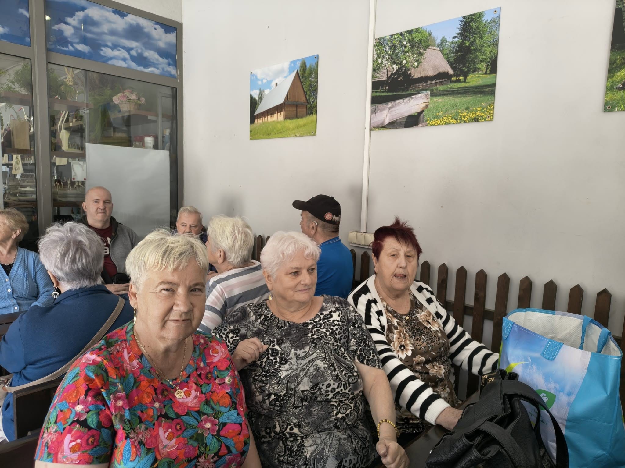 Grupa seniorów odpoczywająca we wnętrzu budynku z obrazami przedstawiającymi wiejskie chaty na ścianach.