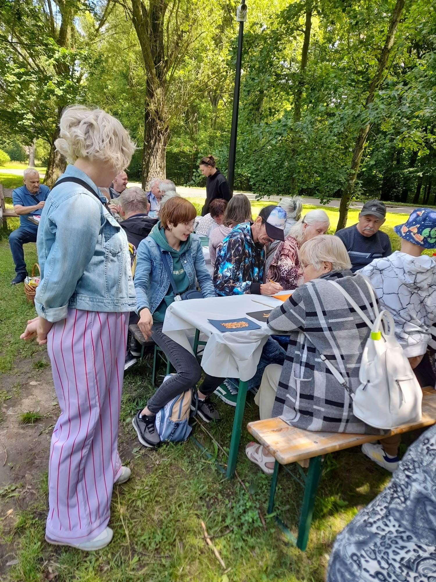 Grupa seniorów oraz opiekunów tworząca prace plastyczne przy stole na świeżym powietrzu w otoczeniu drzew.