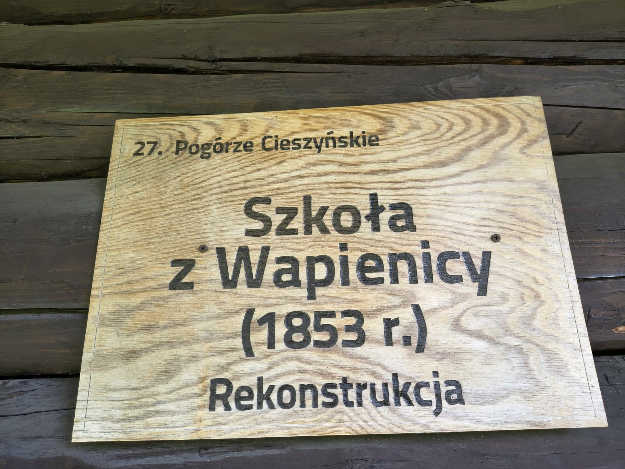 Drewniana tablica informacyjna z napisem „27. Pogórze Cieszyńskie. Szkoła z Wapienicy (1853 r.) Rekonstrukcja”.
