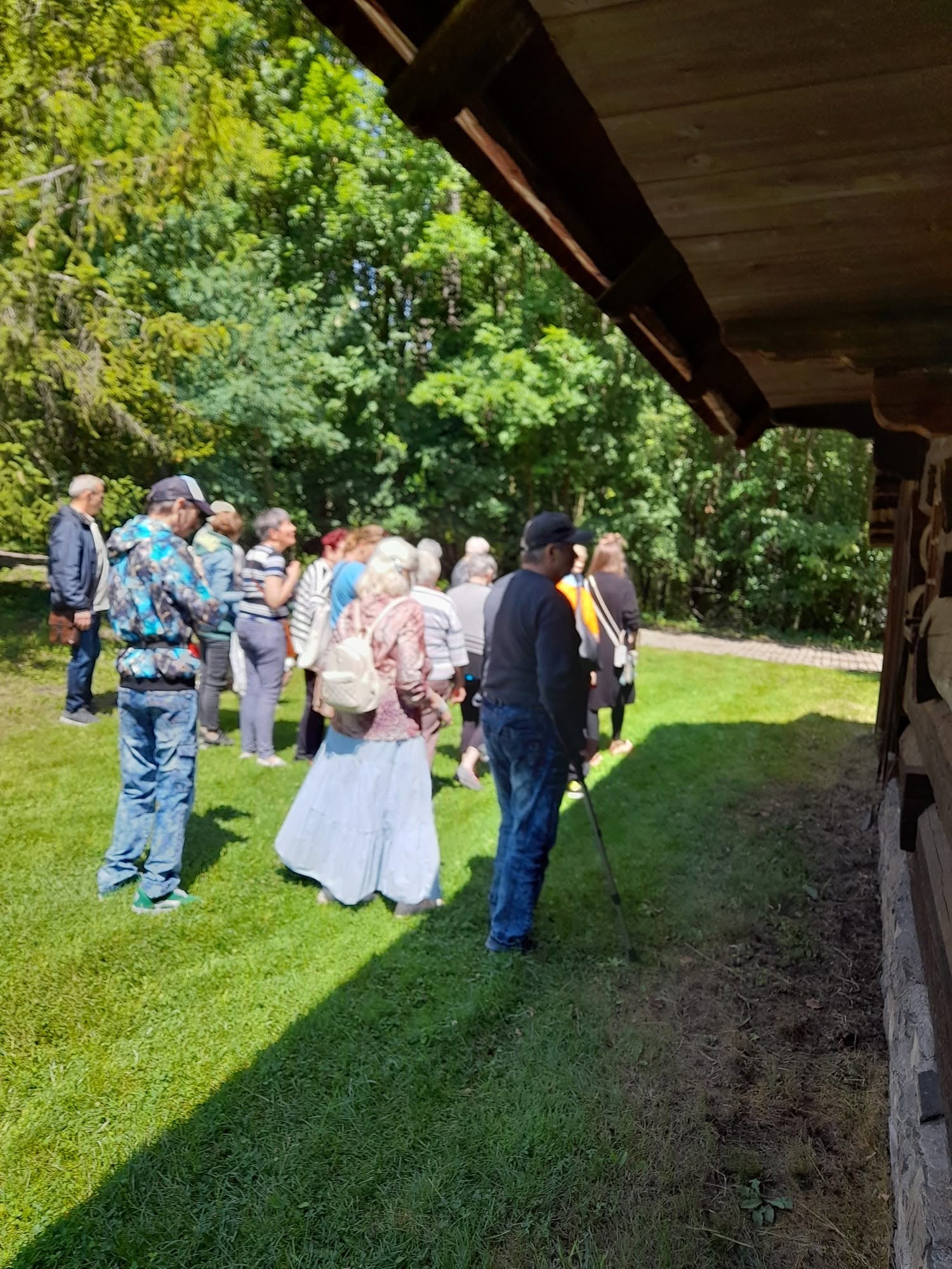 Grupa osób spacerująca w plenerze wzdłuż drewnianego budynku.