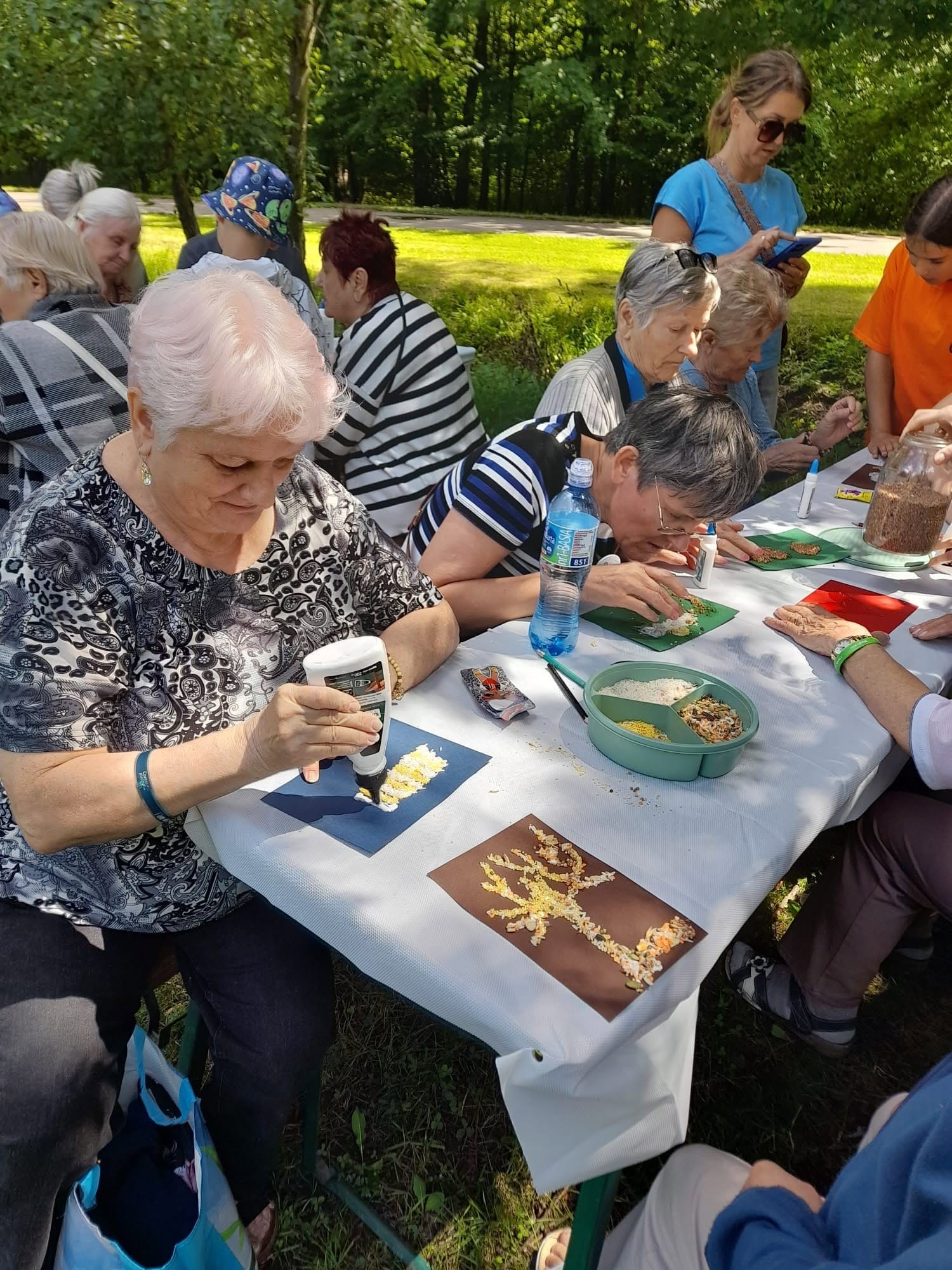 Seniorki uczestniczące w warsztatach plastycznych na świeżym powietrzu, wyklejające mozaiki z ziaren przy stole plenerowym.