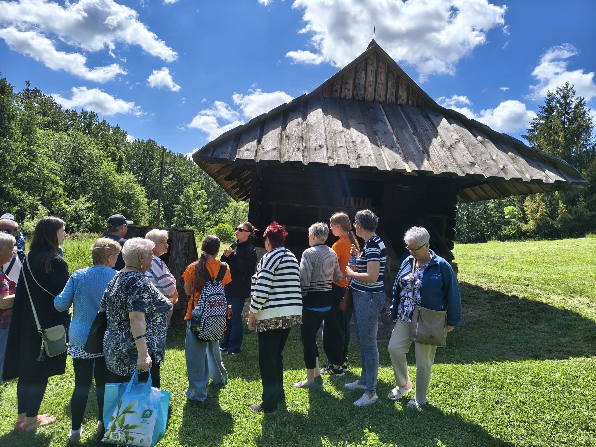 Grupa osób zwiedzająca skansen na świeżym powietrzu, stojąca przed zabytkowym drewnianym budynkiem.