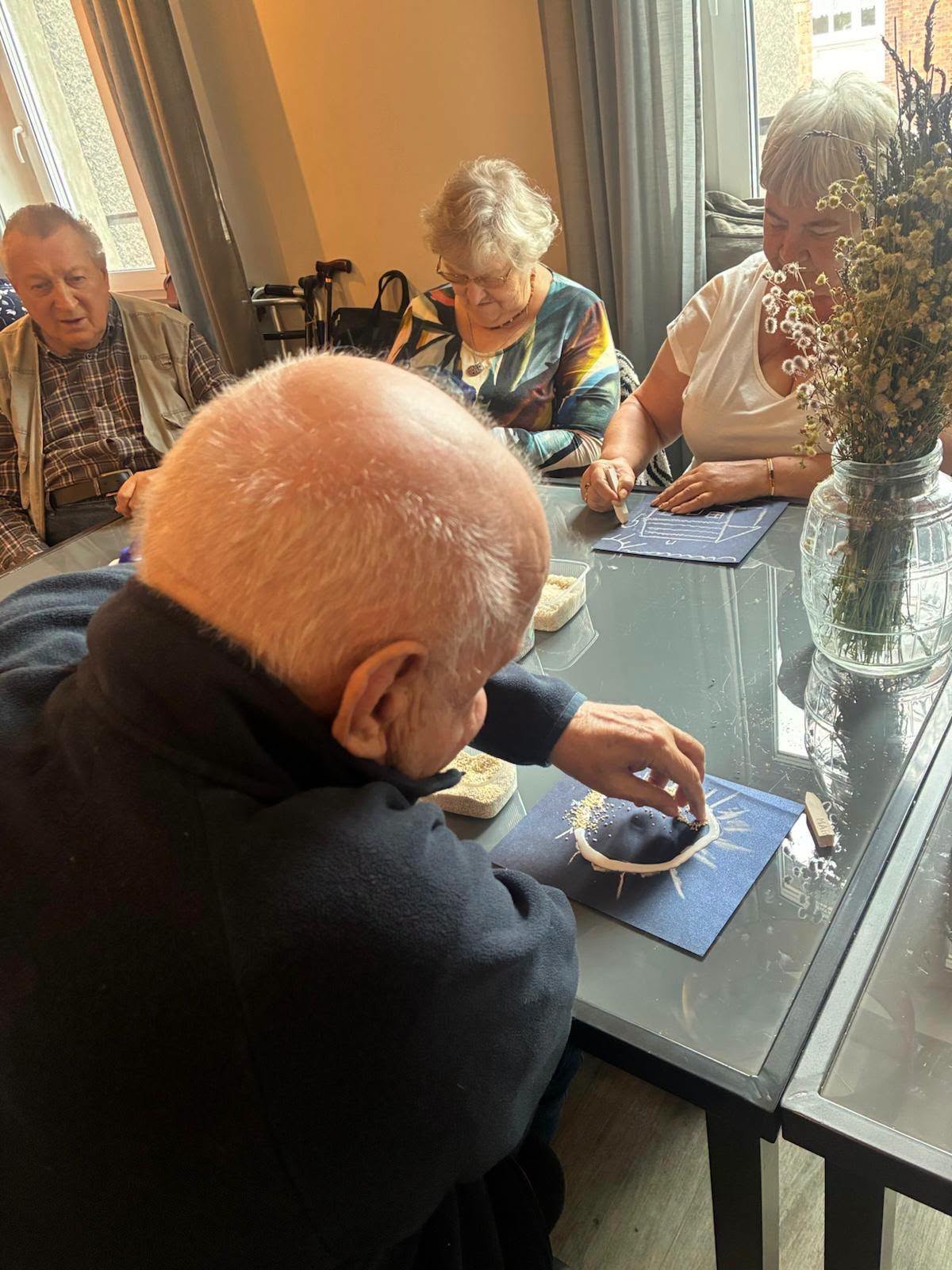 Seniorzy skupieni na tworzeniu prac plastycznych z ziaren przy stole.