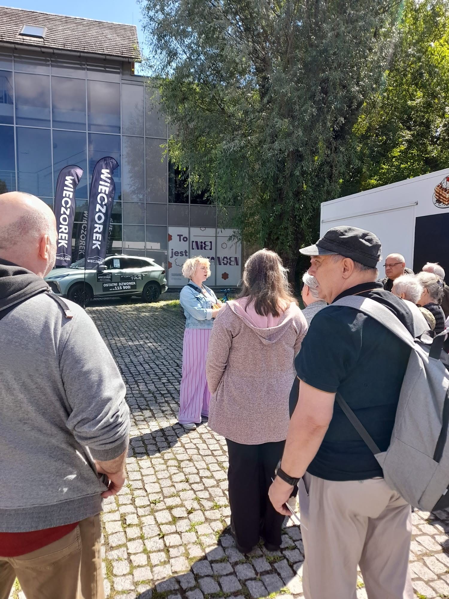 Grupa osób słuchająca przewodnik na placu przed nowoczesnym budynkiem.