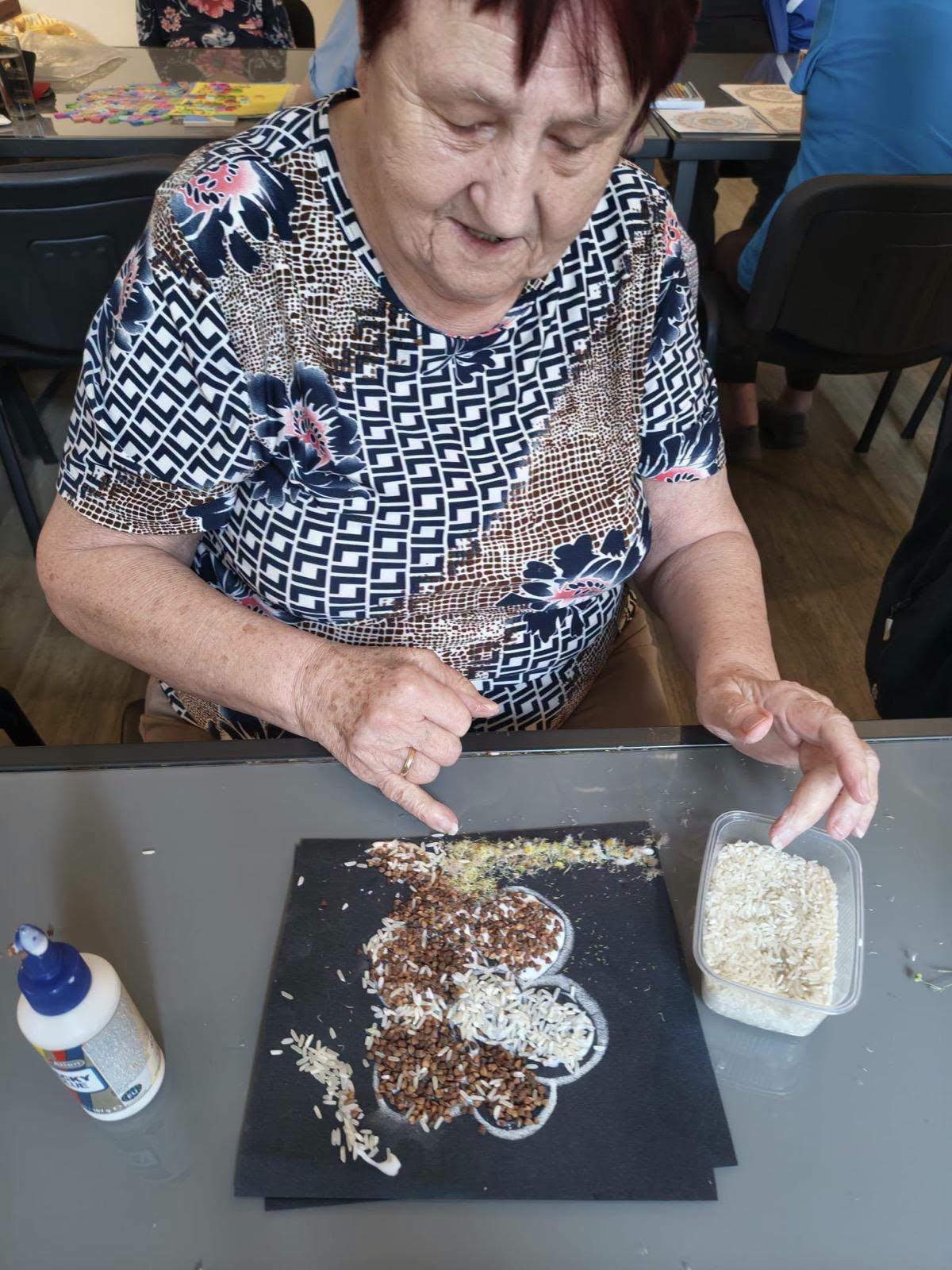 Seniorka wyklejająca kwiatka ziarenkami ryżu i gryki na czarnej kartce.