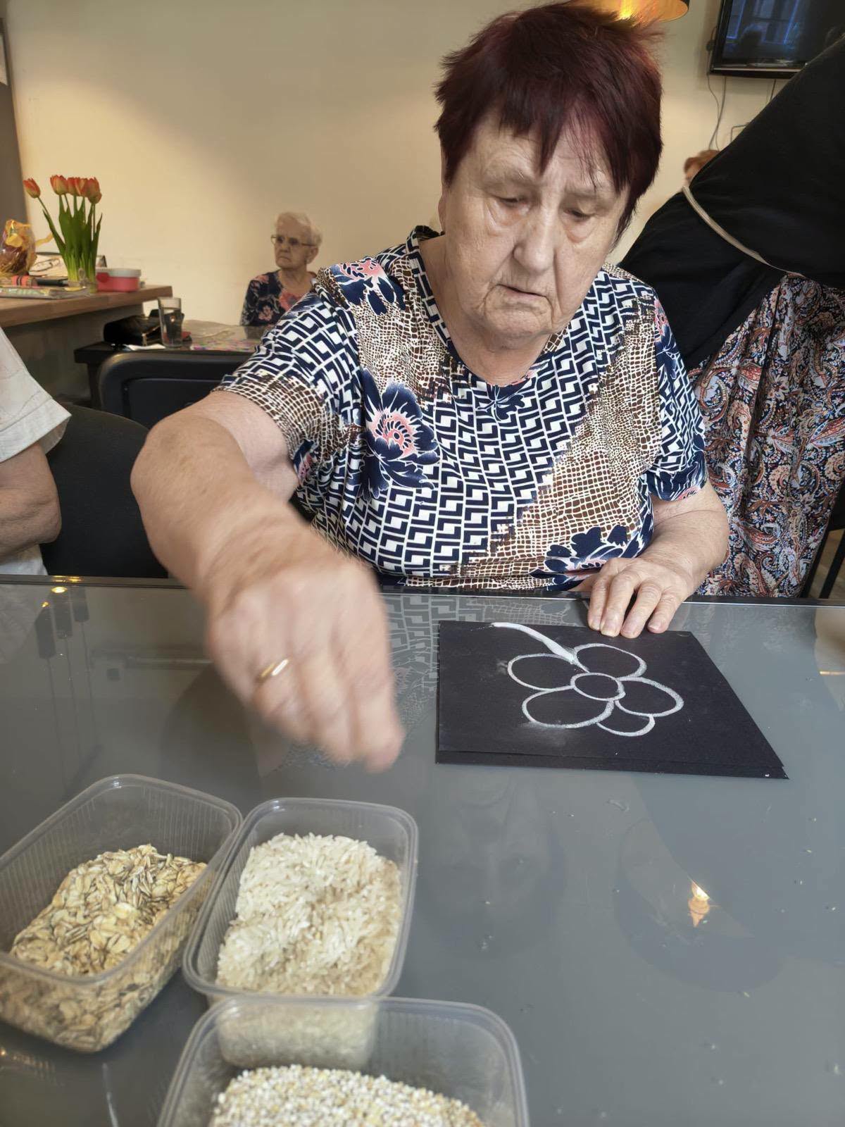 Seniorka układa ziarna na narysowanym kwiatku podczas zajęć plastycznych.