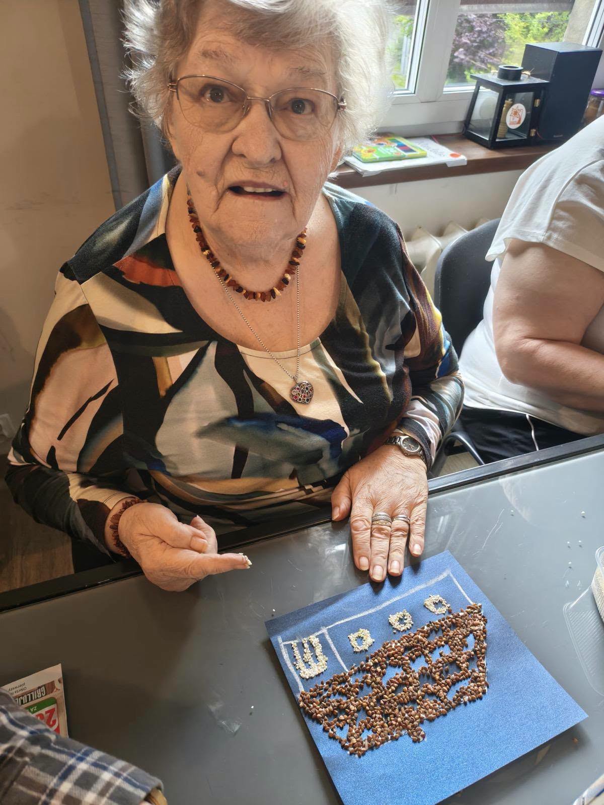Seniorka prezentująca pracę plastyczną z wyklejonym domkiem z gryki.