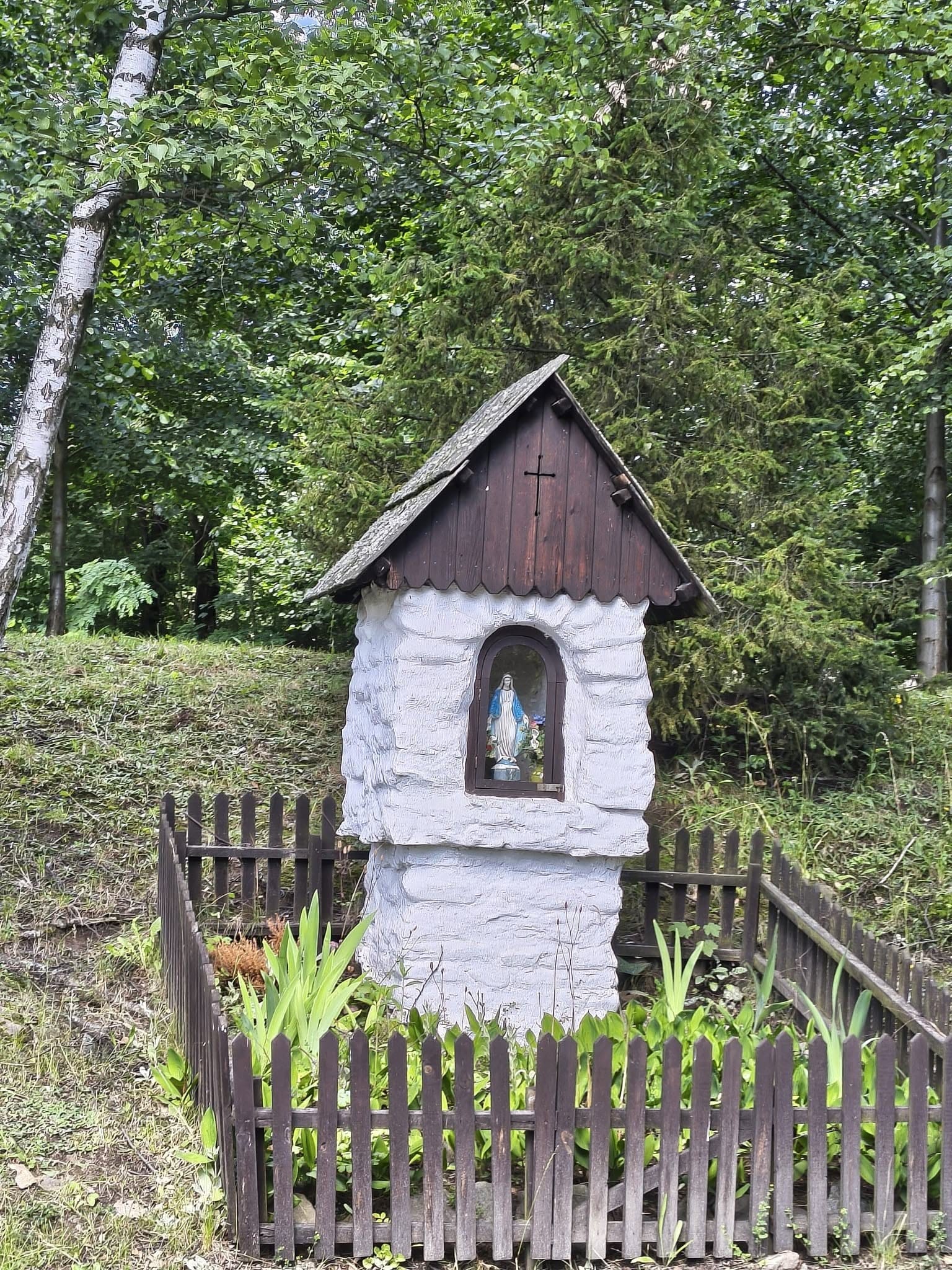 Mała, murowana kapliczka przydrożna z figurką Matki Boskiej we wnęce, otoczona drewnianym płotkiem na tle zieleni.