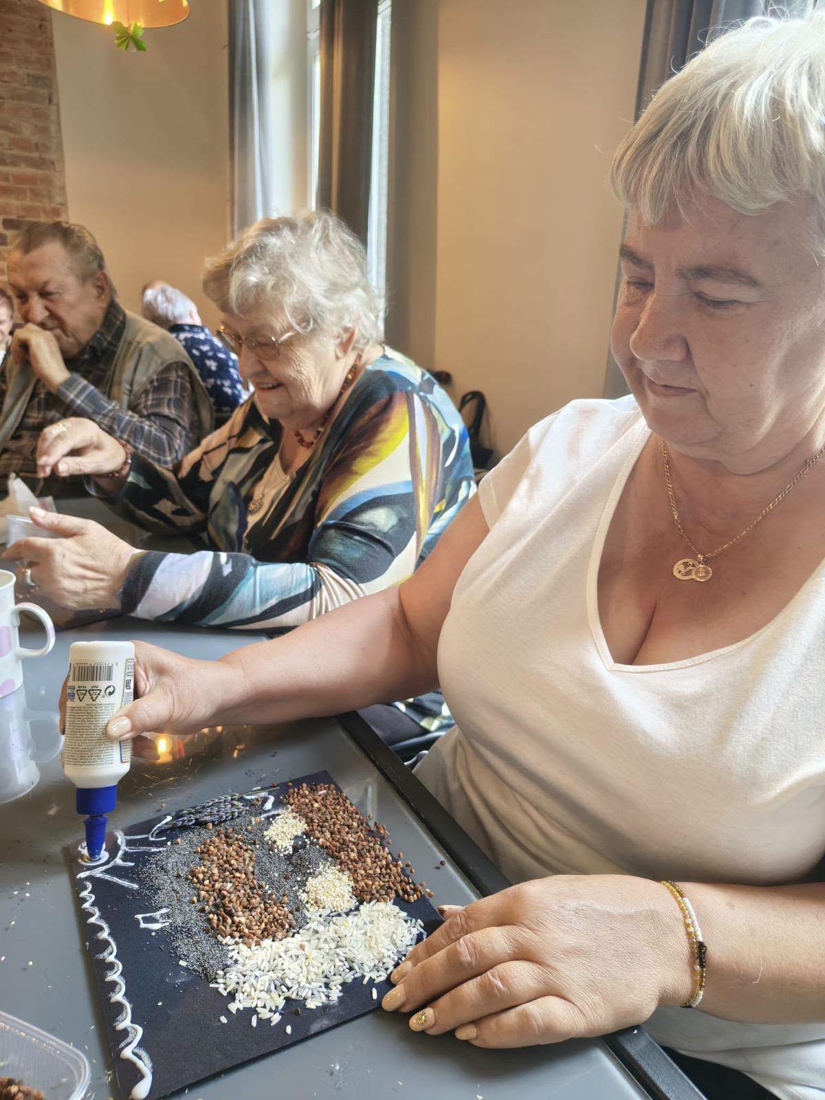Seniorka nakłada klej na mozaikę z nasion i ziaren podczas warsztatów plastycznych.