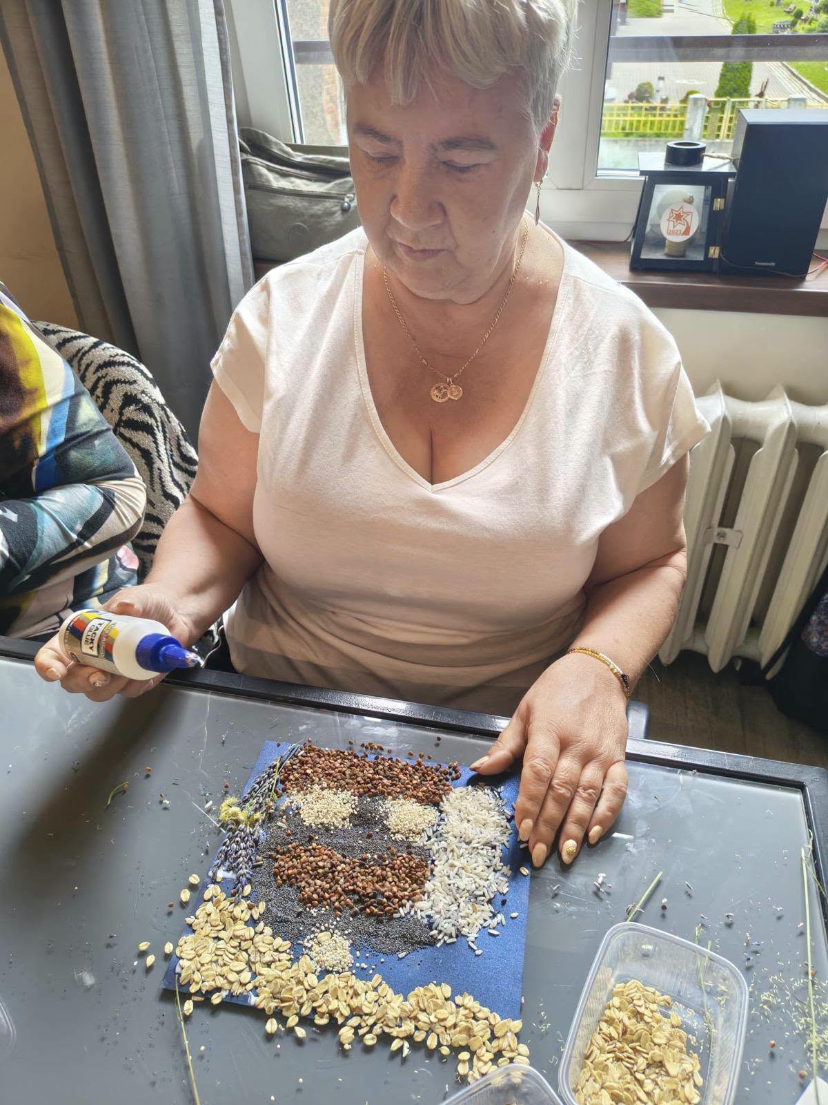 Seniorka trzymająca klej nad tworzoną mozaiką z różnorodnych nasion i ziaren.