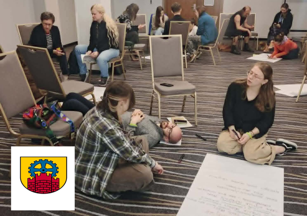Uczestnicy warsztatów pracują w grupach na podłodze i krzesłach, pisząc na arkuszach.
